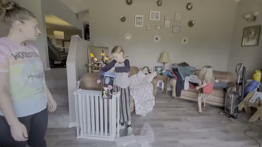 Video of a family living room: a girl in striped pants opening a gift bag by the stair gate, another child in a red shirt watching, a large dog crossing the floor.