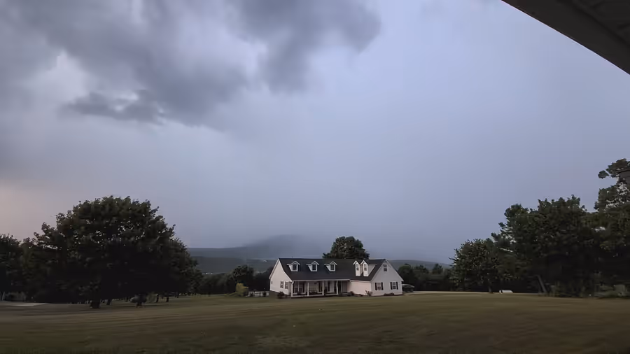 A dark storm front filling the sky above a white farmhouse across a wide lawn, the ridge behind it disappearing into rain.