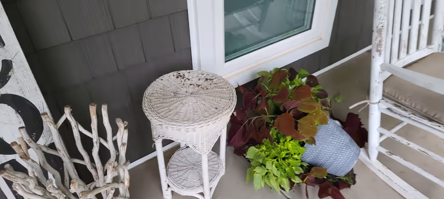 A covered porch with a white wicker table and rocking chair, and a grey planter of purple and green coleus tipped over onto its side on the concrete.