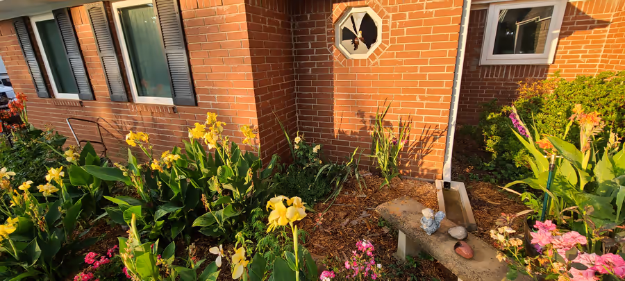 The brick corner of the house in golden evening light, yellow canna lilies and pink flowers in the bed below, the broken octagonal window high on the wall above.
